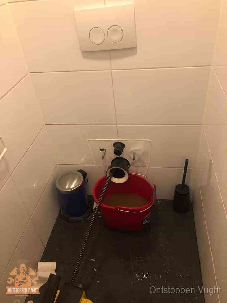 Een ontstopte wand-wc met verwijderde toiletpot waar een ontstoppingsveer in de wanduitgang verdwijnt, het achterhalen waar de verstopping zit in Vught